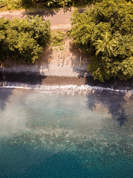 Aerial view of a coastal scene in São Tomé with clear blue waters, a beachfront lined with lush greenery, and shadows of palm trees cast on the sand.