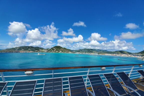 A view from a cruise ship deck overlooking the clear blue waters and the hilly landscape of Sint Maarten under a partly cloudy sky.