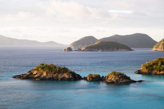 A picturesque view of Trunk Bay, Saint John with turquoise waters, green islands, hills, and a pastel sky.