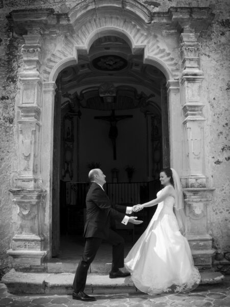 A black and white photo of a bride and groom holding hands in front of an old church entrance in Mexico.