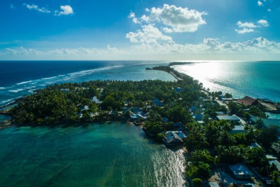 Aerial shot of a thin land strip with lush vegetation and structures, encircled by the Pacific Ocean, possibly Kiribati.