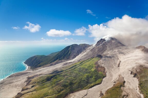 Aerial view shows Soufrière Hills volcano in Montserrat, with smoke, green slopes, and a rugged coastline.