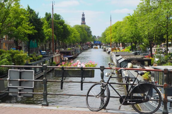 A canal in Amsterdam, Netherlands, with a bicycle in the foreground, trees lining the sides, and a church tower in the distance.