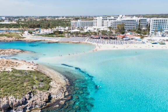 Aerial view of a coastal area in Ayia Napa, Cyprus, featuring clear turquoise waters, sandy beaches, and clusters of white buildings along the shore.