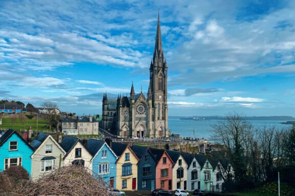 A picturesque view of Cobh, Ireland, showcasing vibrant houses, a towering cathedral, and a partly cloudy sky.