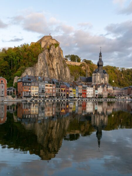 A scenic view of Dinant, Belgium, with vibrant riverside buildings, a church, a cliff, all mirrored in the tranquil river.