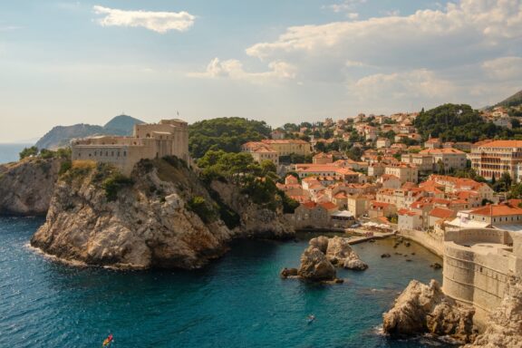 A coastal view of Dubrovnik, Croatia, featuring historic buildings and walls on a cliff overlooking the Adriatic Sea.