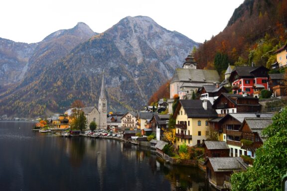 A serene lake, alpine buildings, a church spire, and autumn mountains form a picturesque view of Hallstatt, Austria.