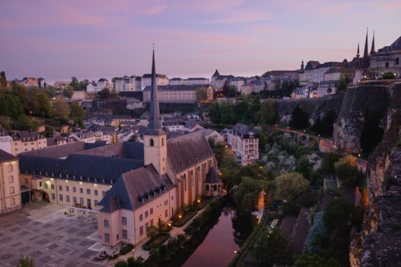 A twilight scene of Luxembourg City, showcasing historical buildings, a river, and trees, softly lit by ambient light.