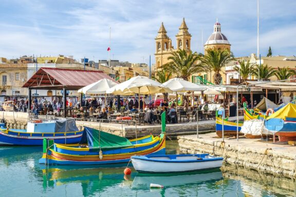 A lively waterfront at Marsaxlokk, Malta with colorful boats, a busy market, people, historical buildings, and church spires under a blue sky.