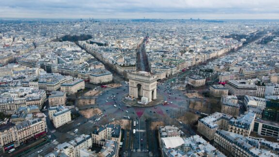 Aerial view of Paris shows the Arc de Triomphe at the center of twelve star-shaped avenues, with cityscape in the background.