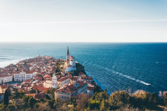 Aerial view of Piran, Slovenia, showcasing the coastal town with red-roofed buildings and a prominent church spire, adjacent to the calm blue sea.