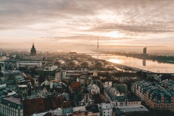 Aerial view of Riga, Latvia during sunrise with mist over the city, showcasing the river, bridges, and a mix of historic and modern buildings.