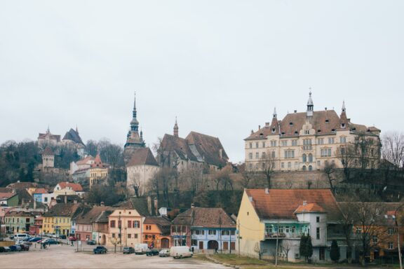 A picturesque view of Sighisoara, Romania, showcasing vibrant houses, a historic hilltop citadel, and a cloudy sky.