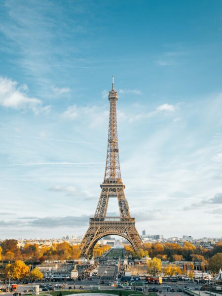 A photograph of the Eiffel Tower in Paris, France, with a clear blue sky in the background and autumn-colored trees below.