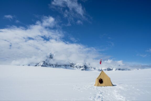 A lone yellow tent stands on a vast expanse of snow with a backdrop of distant mountains under a partly cloudy sky in Antarctica.