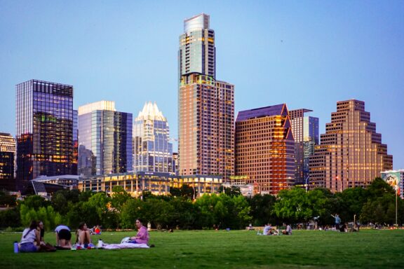 A cityscape of Austin, Texas, with modern high-rise buildings during twilight, and people relaxing on a grassy park in the foreground.