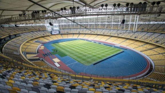 Interior view of the Bukit Jalil Stadium with empty yellow and blue seats, an athletic track, and a green soccer field.