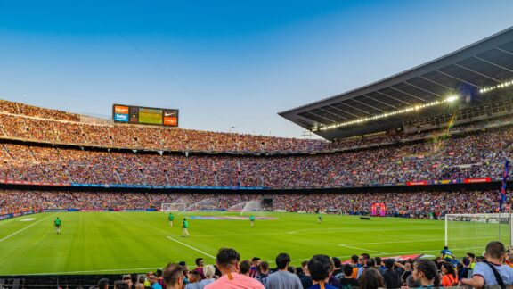 A panoramic view of Camp Nou stadium during a soccer match with spectators in the stands and players on the field at dusk.