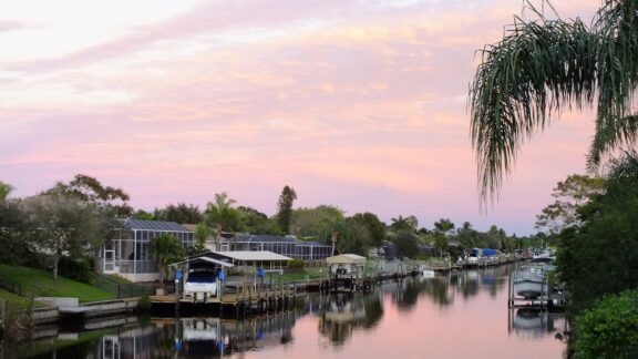 A peaceful sunset in Cape Coral, Florida, featuring a canal, homes, boats, and a foreground palm tree.