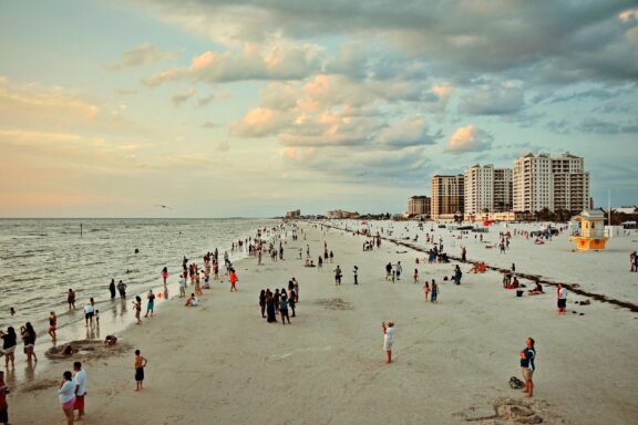 A busy beach in Clearwater, Florida with people enjoying the sand and tall buildings under a cloudy sky.