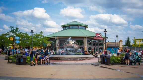 A pavilion with a fountain in front at the Columbus Zoo, with people sitting and walking around on a sunny day with scattered clouds.