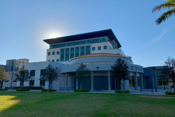 A modern 'CITY HALL' building with a curved facade in Coral Springs, partially obscuring the sun.