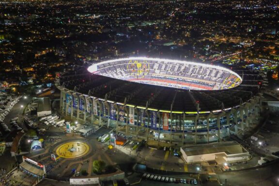 Aerial night view of Estadio Azteca, a large stadium with bright lights, surrounded by a sprawling cityscape.