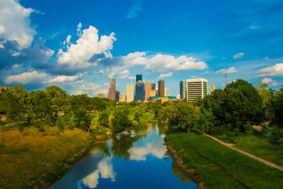 A picturesque view of Houston, Texas, with a river, lush greenery, city skyline, and a blue sky with clouds.