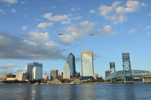 A view of the Jacksonville, Florida skyline with buildings reflected in the water, a bridge to the right, and a clear sky with clouds above.