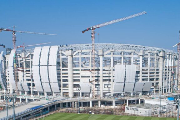 An image showing the construction of the Jakarta International Stadium with cranes and a clear blue sky in the background.