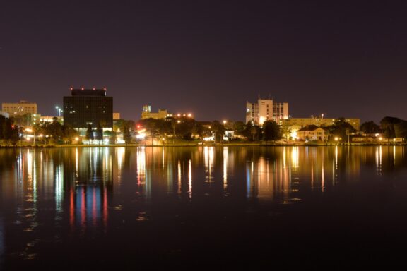 A nighttime cityscape of Lakeland, Florida, with illuminated buildings reflected in a calm lake.