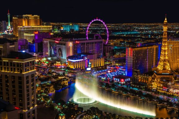 A nighttime view of the Las Vegas Strip in Nevada, showcasing illuminated hotels, a large observation wheel, and a fountain show in the foreground.