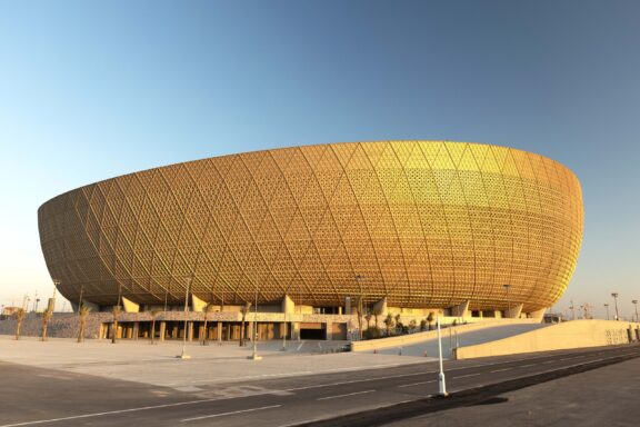 A photo displays the unique gold, rounded facade of Lusail Stadium in Qatar under a clear sky at sunrise or sunset.