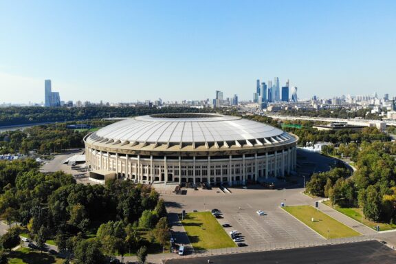 Aerial view of Luzhniki Stadium in Moscow with city skyline in the background, clear skies, and surrounding greenery.