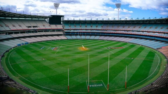 An empty Melbourne Cricket Ground stadium with green grass and seating visible under a partly cloudy sky.