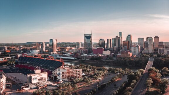 Aerial view of downtown Nashville, Tennessee, with a clear sky, showcasing the city's skyline and buildings, including a stadium in the foreground.