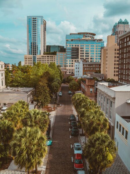 Aerial view of a tree-lined street in downtown Orlando, Florida, with tall buildings on either side under a partly cloudy sky.