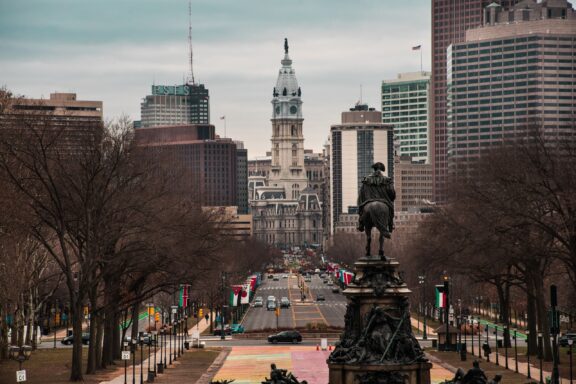 A city street in Philadelphia leads to a historic clock tower, surrounded by skyscrapers and a statue under a cloudy sky.