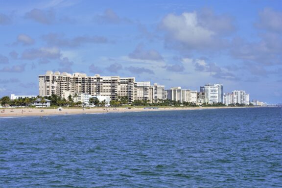 A scenic coastal view of Pompano Beach featuring a sandy shore, high-rise buildings, and a blue sky with clouds.