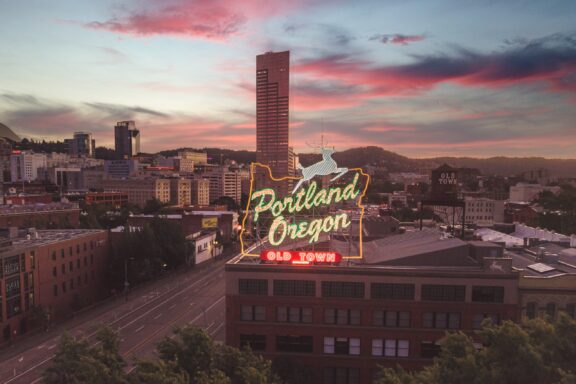 Aerial view of downtown Portland, Oregon at dusk with buildings, a neon sign reading "Portland Oregon Old Town," and a colorful sky.