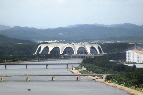 A distant view of the large, domed Rungrado 1st of May Stadium, located near a river and mountains under a cloudy sky.