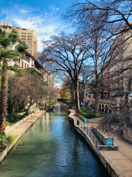 A scenic view of the San Antonio Riverwalk in Texas, featuring a calm river flanked by walkways, trees, and buildings under a clear blue sky.