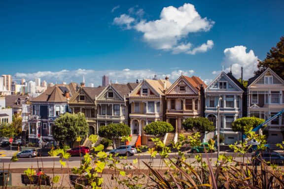 A row of Victorian houses in San Francisco, California, with a clear blue sky and some city buildings in the background.