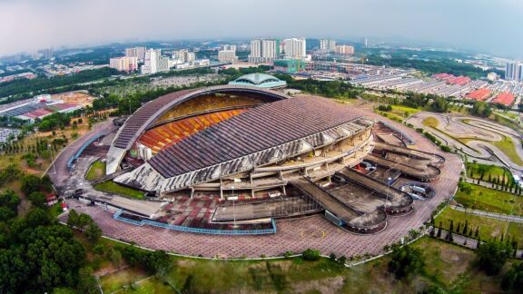 Aerial view of Shah Alam Stadium with its distinctive architecture, surrounded by parking areas and greenery, under a hazy sky.