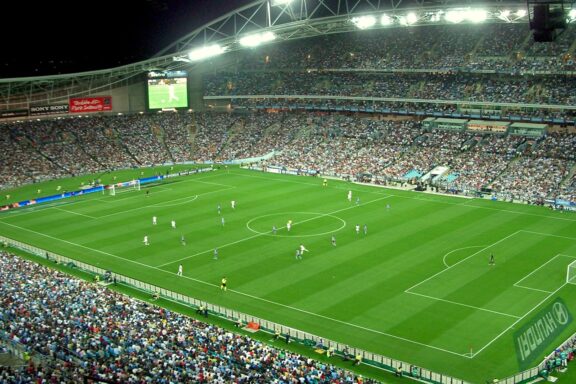 A nighttime event at Stadium Australia, showcasing a lit soccer field, active players, and a large audience.