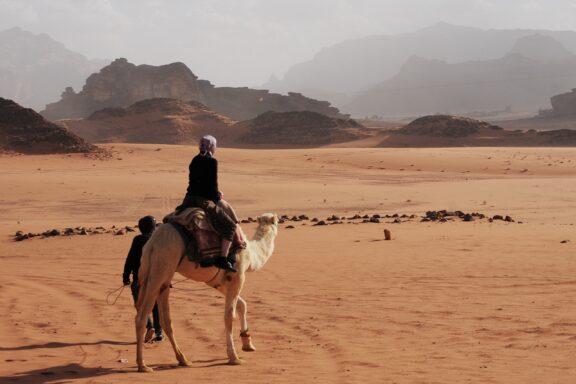 A person riding a camel in the Wadi Rum desert, Jordan, with rocky mountains in the background and a vast sandy landscape.