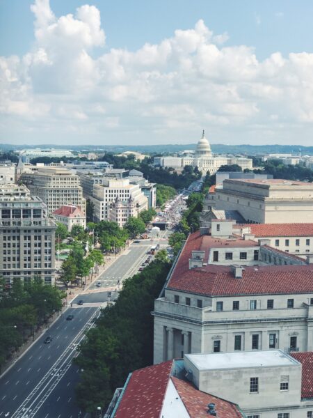 Aerial view of Washington DC with the Capitol building in the distance, streets, and buildings in the foreground under a partly cloudy sky.