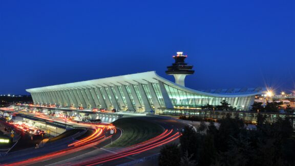 Night view of Washington Dulles International Airport with illuminated terminal building and control tower, traffic streaks visible.