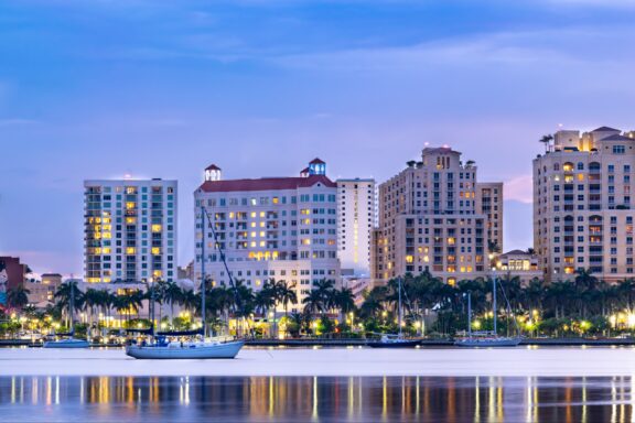 A twilight view of the waterfront skyline of West Palm Beach, Florida, with reflections on the water's surface.